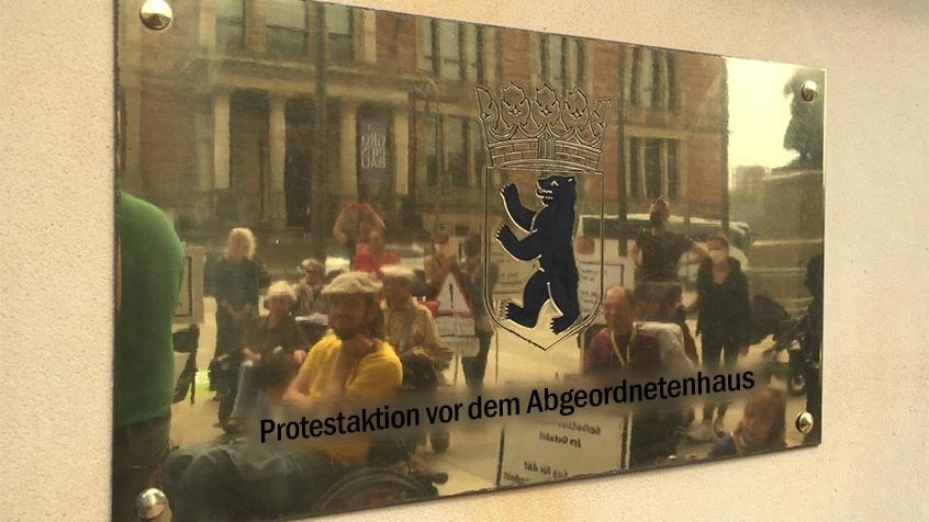 Rechteckiges gold glänzendes Schild mit dem Wappen Berlins in der Mitte.
Gespiegelt in dem Schild sind ca. 14 Menschen mit und ohne Rollstuhl, teilweise mit Protestschildern zu sehen.
Unter dem Wappen ein eingefügter Schriftzug: Protestaktion vor dem Abgeordnetenhaus
